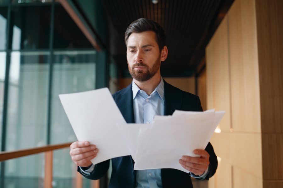 Executive examining documents feeling excitement walking office hallway. Emotional businessman throwing papers rejoicing business success. Overjoyed manager winning deal gesturing joyfully at corridor